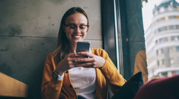 A woman smiling and looking at her cell phone while leaning against a wall