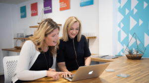 Two smilng adult woman using a laptop while sitting in an office space