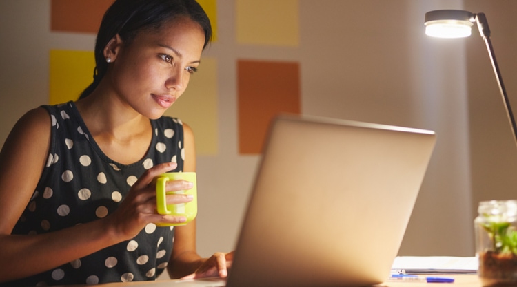 A woman holding a mug in an office while typing on a laptop