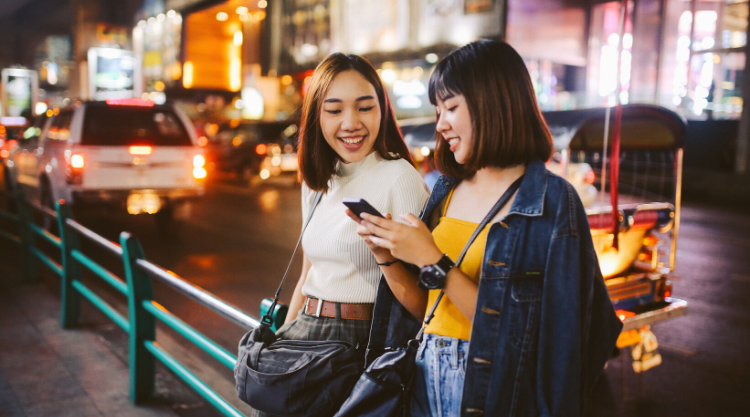 Two smiling young woman leaning against a railing in front of a road while looking at one of their cellphones