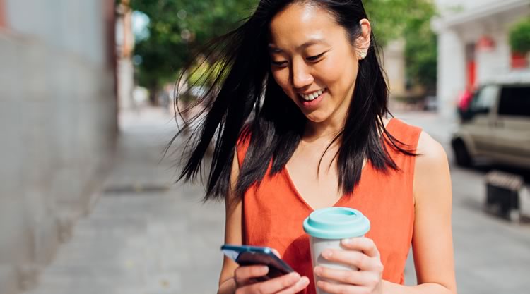 A smiling woman looks down at her phone while holding a cup of coffee in a parking lot.