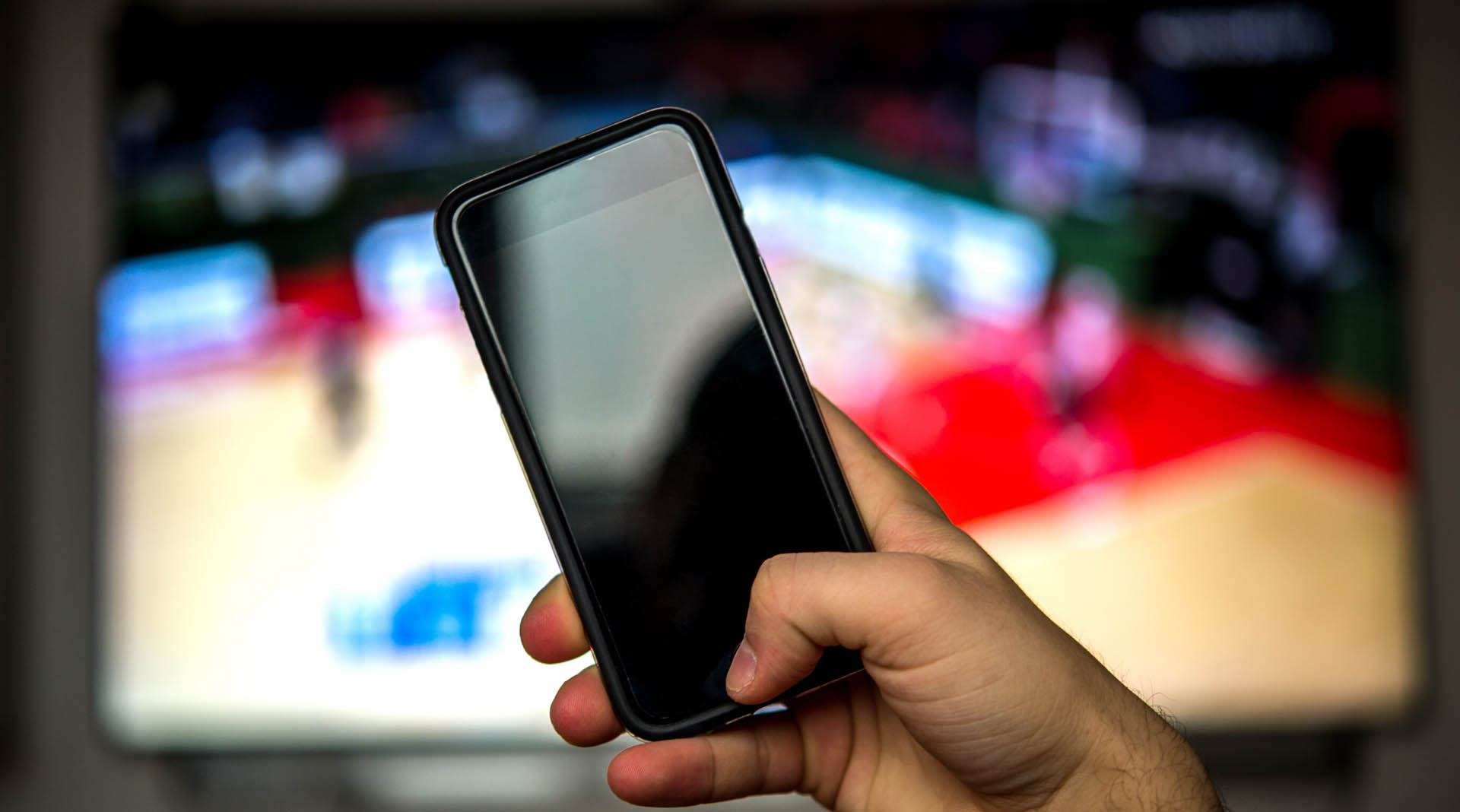 A hand holding a cell phone with the a television blurred in the background