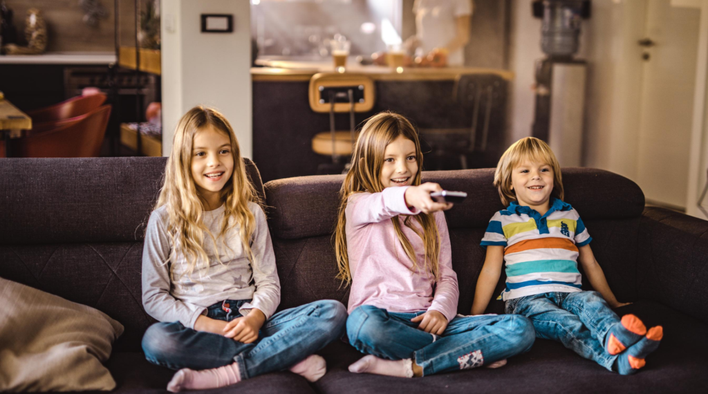 Three children sitting on their couch in their living room. The child in the center is holding a TV remote.