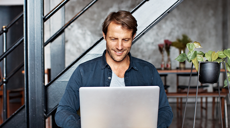 A smiling man seated and in an office space working on his laptop