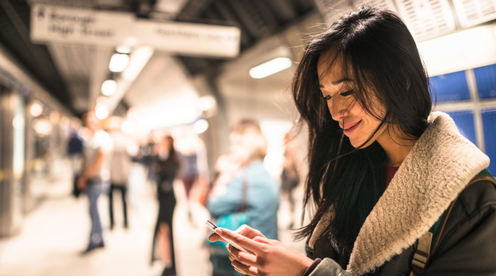 A young smiling woman looks down at her phone while texting at a train station.