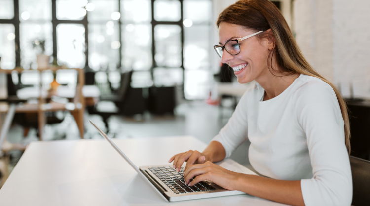 A smiling woman seated in an open office space and is working on her laptop