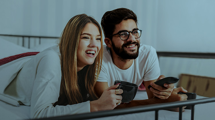 Young woman smiling holding a mug laying in bed next to man holding a remote