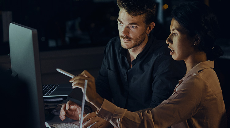 Adult woman point at a computer screen with a pen in hand while an adult man looks at the screen in an office space