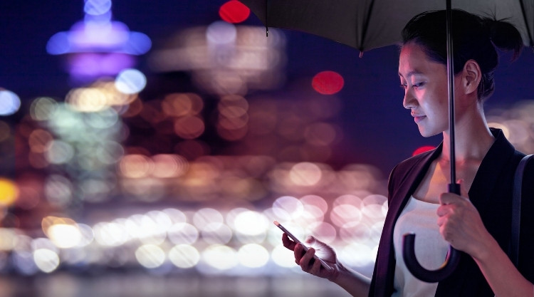A smiling woman holding an umbrella looks down at her cellphone in hand, there's a blurred view of a city behind her.