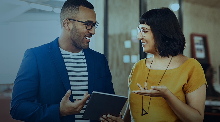 Smiling adult man holding a tablet and looking at a smiling adult woman while in an office space