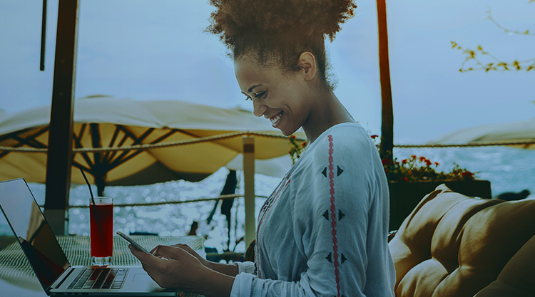 A smiling adult woman sitting by the water with a glass of juice and laptop open in front of her. She also has a cell phone in hand