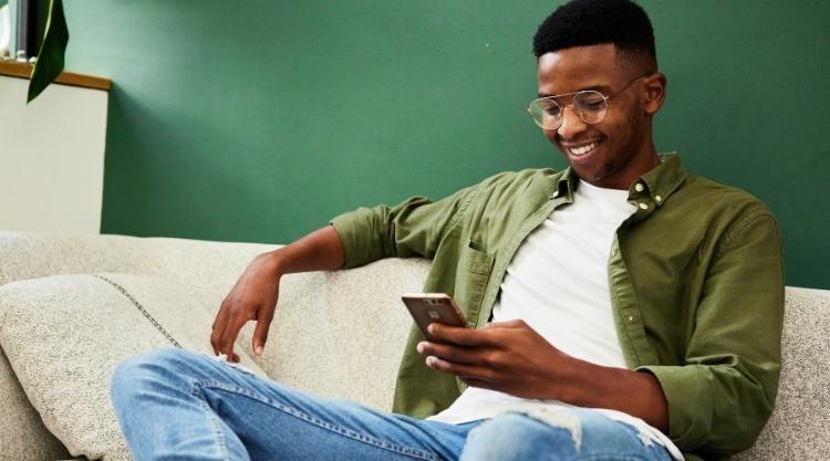 Young African man smiling at a text massage on his mobile phone while relaxing on his living room sofa at home