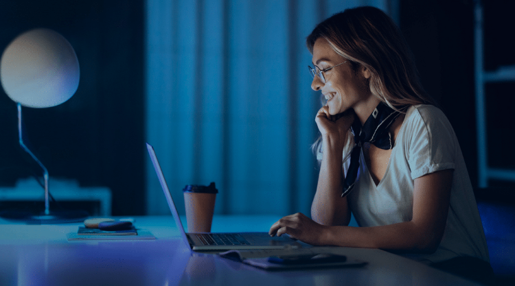 A smiling adult woman typing on her laptop in a dark room with blue lighting